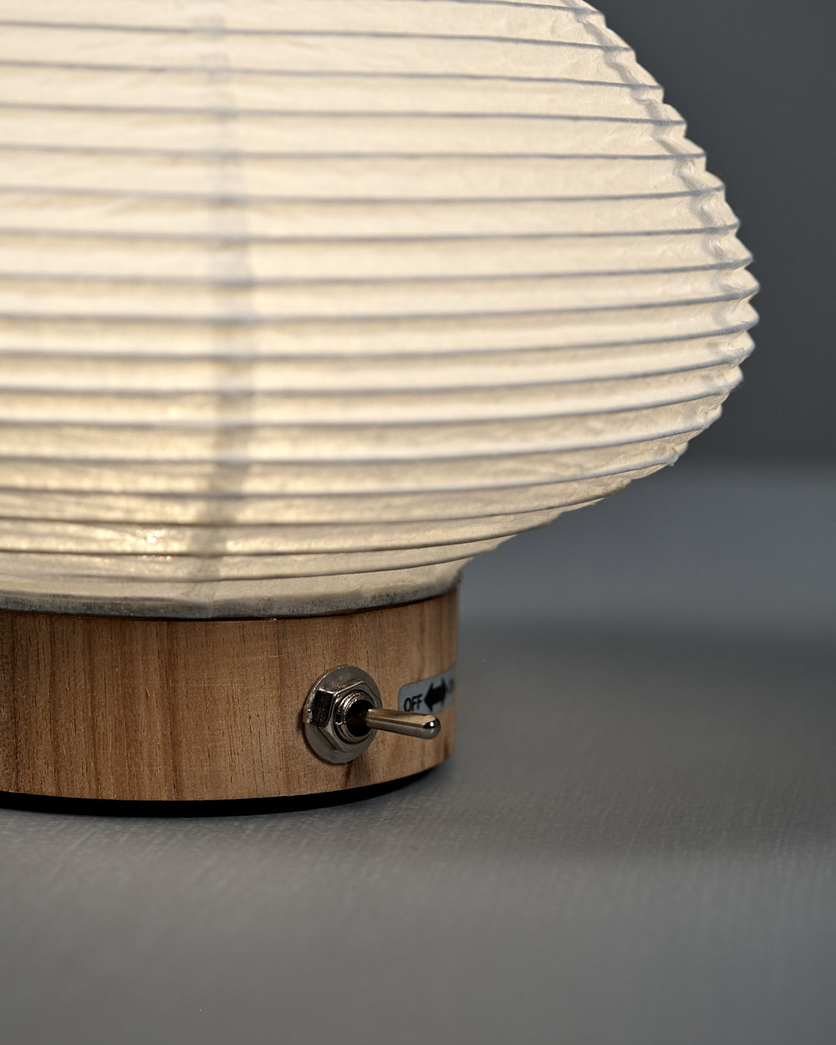 A close-up of the Washi Paper LED Lantern – Musubi, featuring a wooden base, white textured shade, and metal toggle switch labeled “off,” crafted by Japanese designer Rina Ono for Rinao design studio.