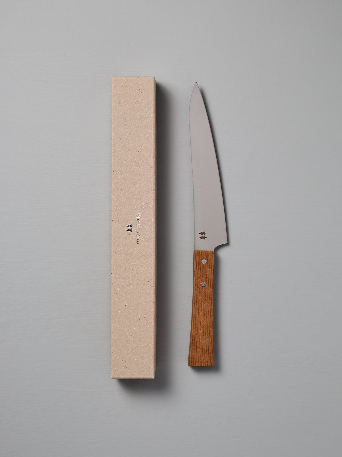 The Morinoki Ban Nou Knife by Shizu Hamono Co., featuring an ergonomic handle and razor-sharp blade, lies next to its beige rectangular case against a gray background.