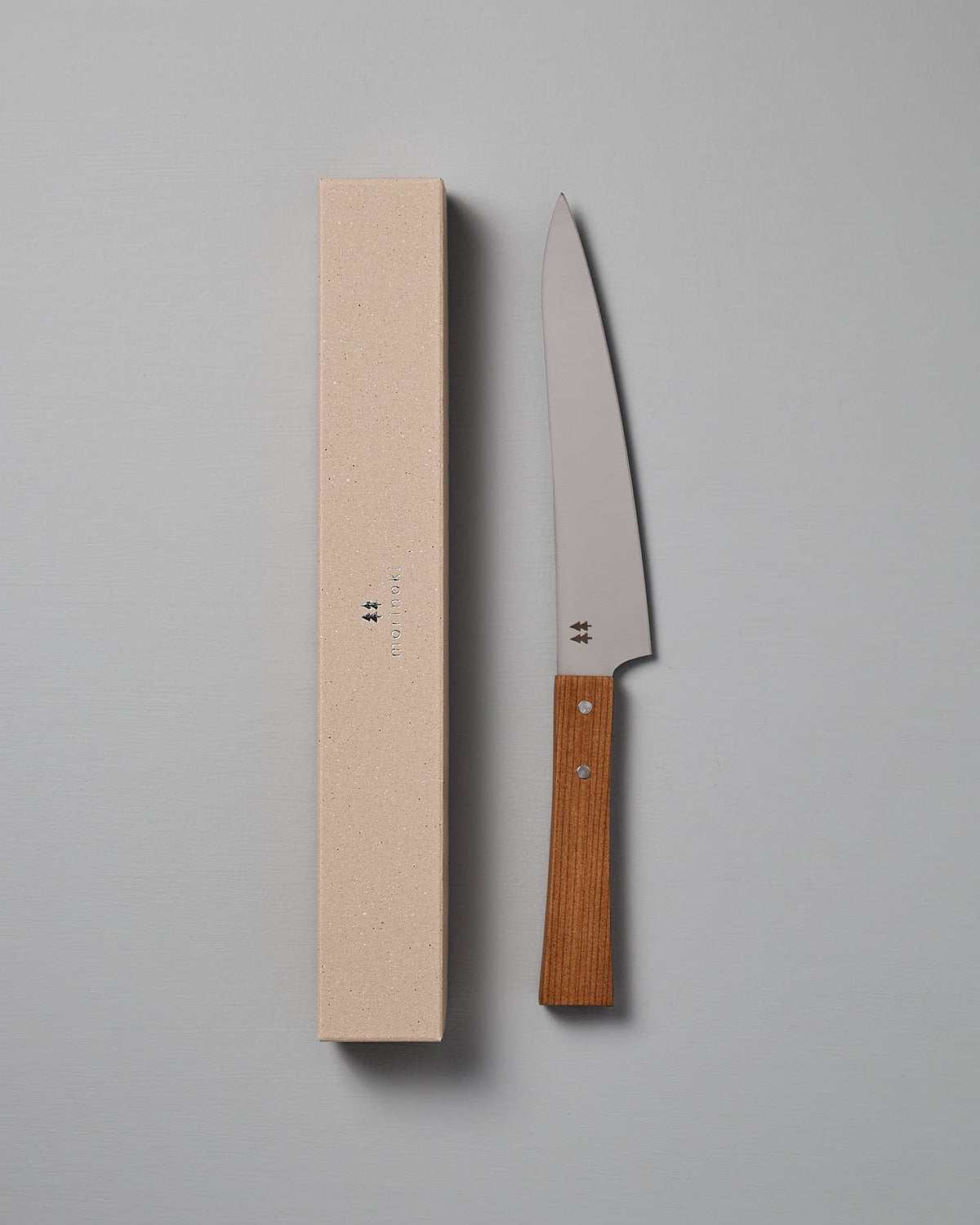 The Morinoki Ban Nou Knife by Shizu Hamono Co., featuring an ergonomic handle and razor-sharp blade, lies next to its beige rectangular case against a gray background.