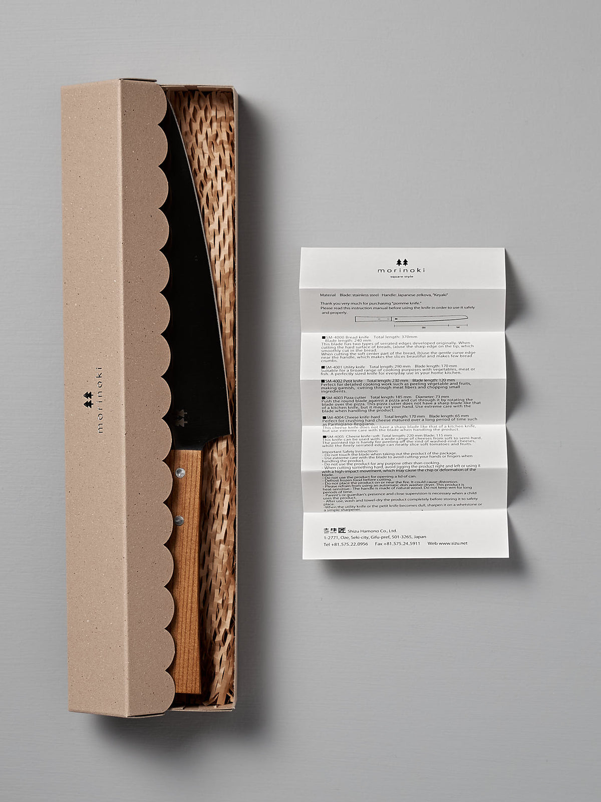 A Morinoki Ban Nou Knife by Shizu Hamono Co. features a razor-sharp blade and an ergonomic handle, partially enclosed in a cardboard box with a wicker-like interior. Next to the box, on a gray surface, lies an unfolded instruction paper.