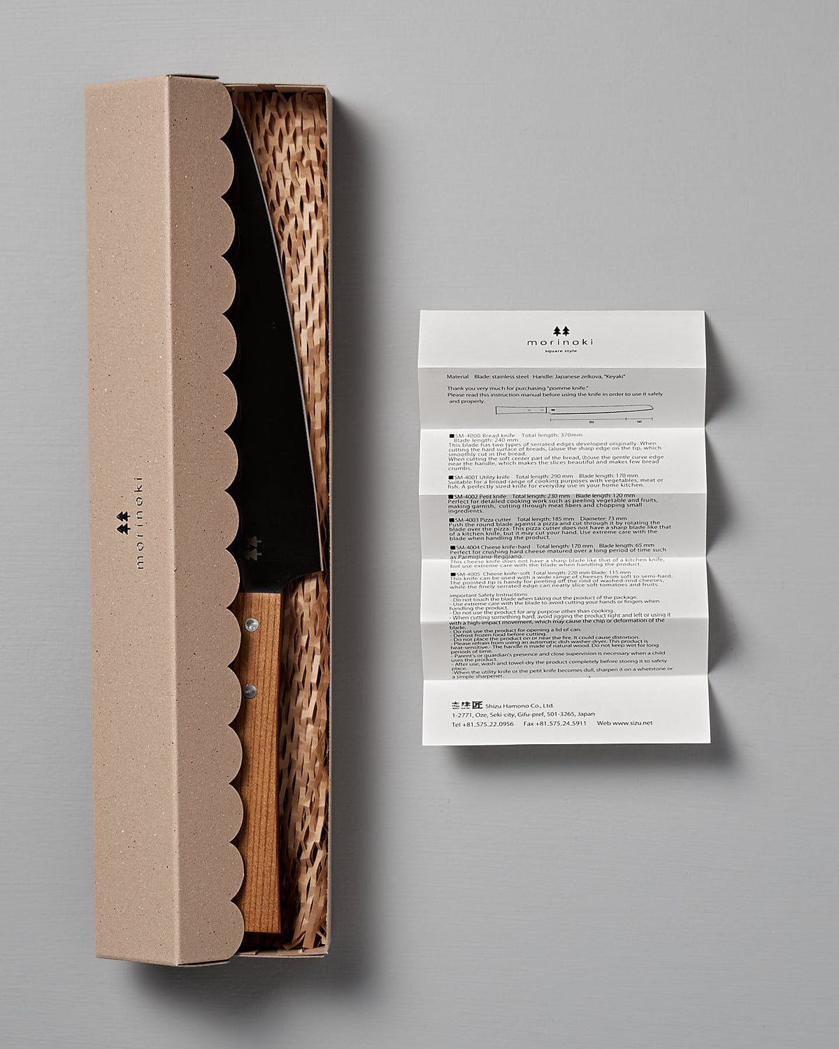 A Morinoki Ban Nou Knife by Shizu Hamono Co. features a razor-sharp blade and an ergonomic handle, partially enclosed in a cardboard box with a wicker-like interior. Next to the box, on a gray surface, lies an unfolded instruction paper.
