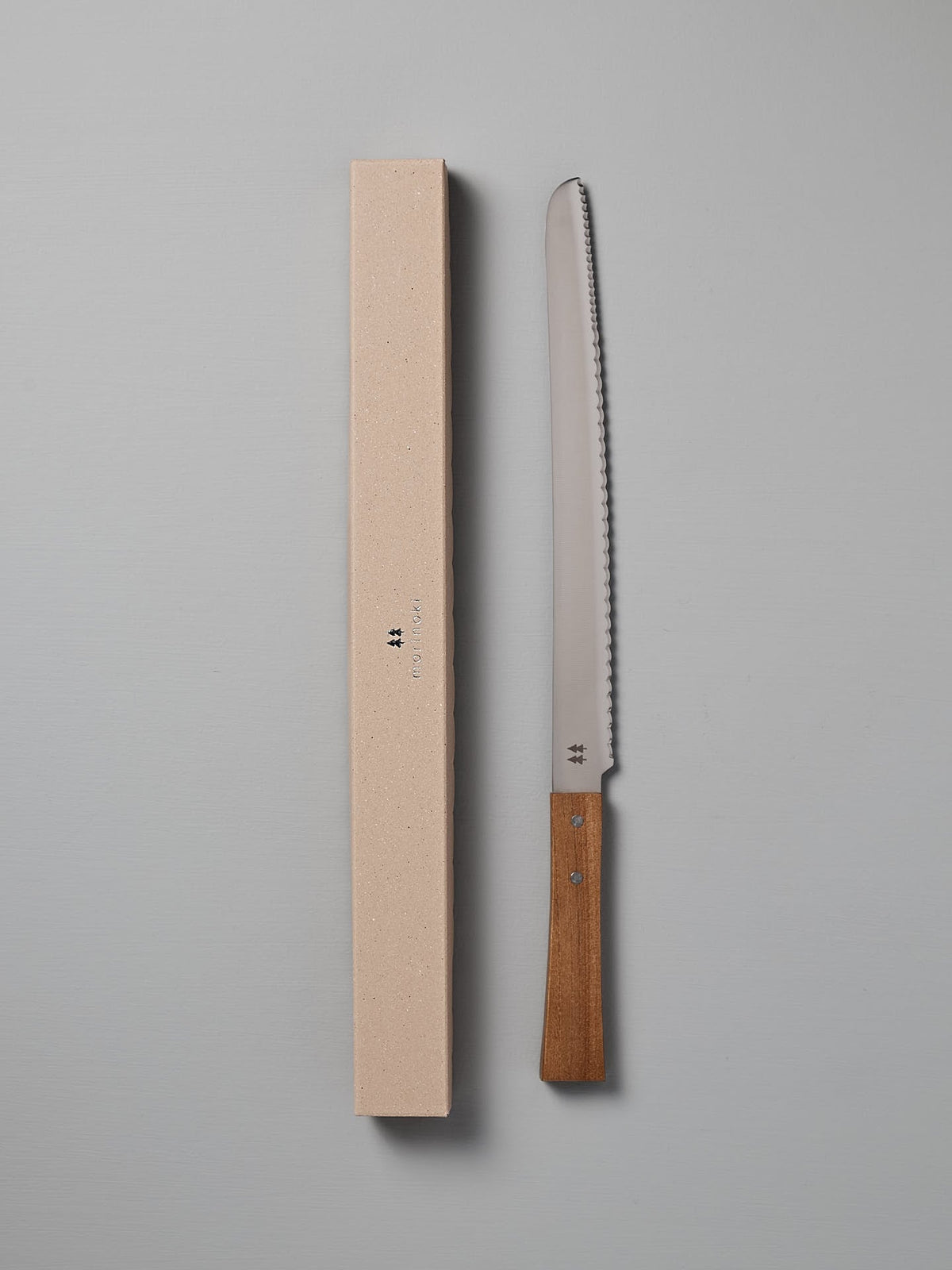 A Morinoki Bread Knife by Shizu Hamono Co., featuring a serrated stainless steel blade and a handle made from Japanese Zelkova wood, rests beside its beige, rectangular box on a gray surface.