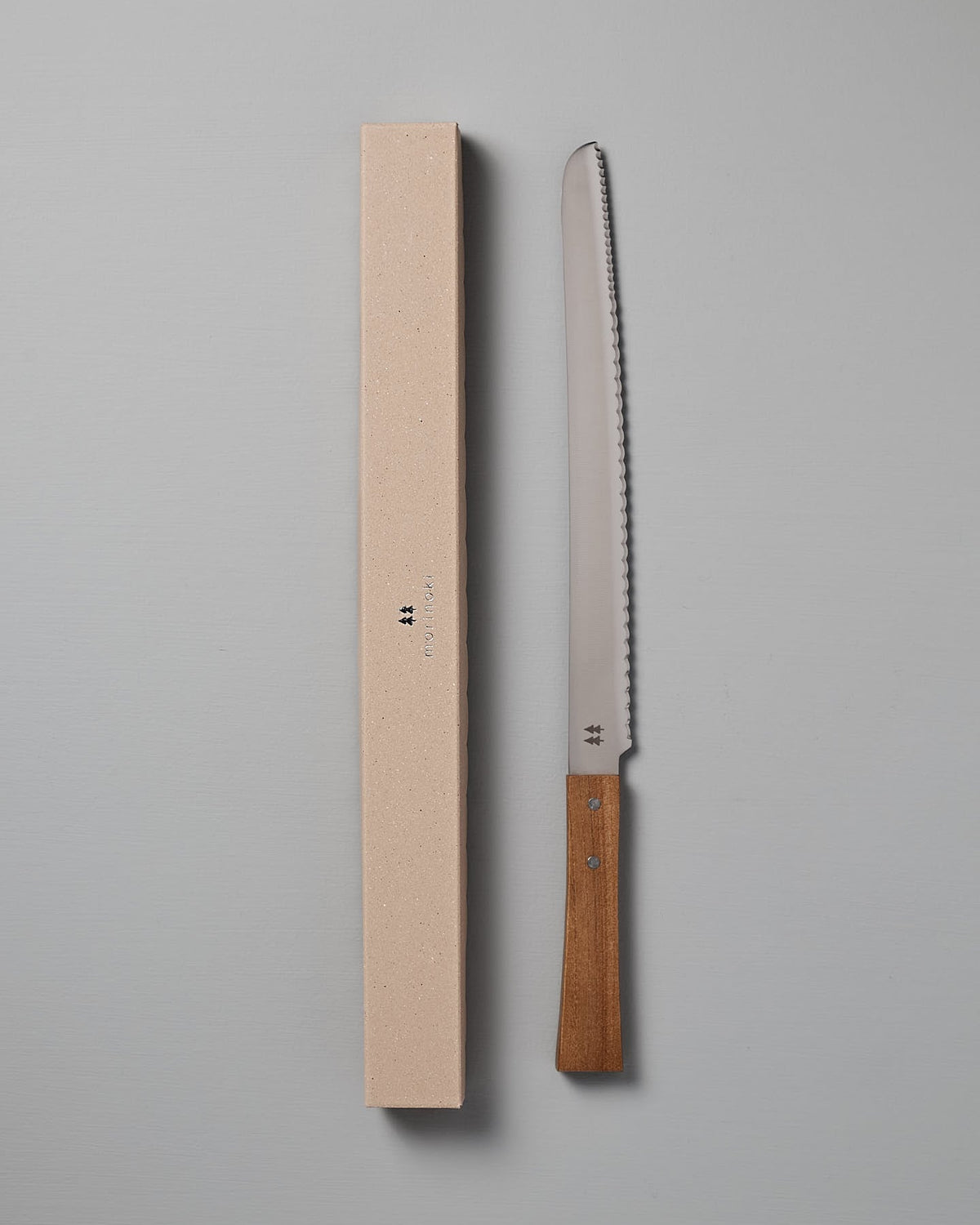 A Morinoki Bread Knife by Shizu Hamono Co., featuring a serrated stainless steel blade and a handle made from Japanese Zelkova wood, rests beside its beige, rectangular box on a gray surface.