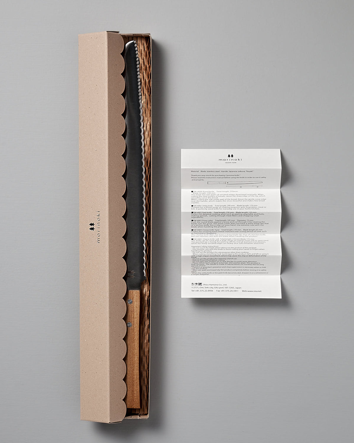 A Shizu Hamono Co. Morinoki Bread Knife, featuring a serrated stainless steel blade and a Japanese Zelkova wood handle, rests in an open cardboard box with a wavy edge design. To the right, a folded instruction manual is laid out flat on a gray surface.