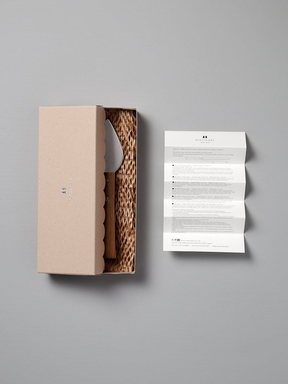 A beige box with scalloped edges holds the Morinoki Hard Cheese Knife by Shizu Hamono Co., featuring a stainless steel blade on textured padding, beside a folded instruction sheet on a light gray surface.