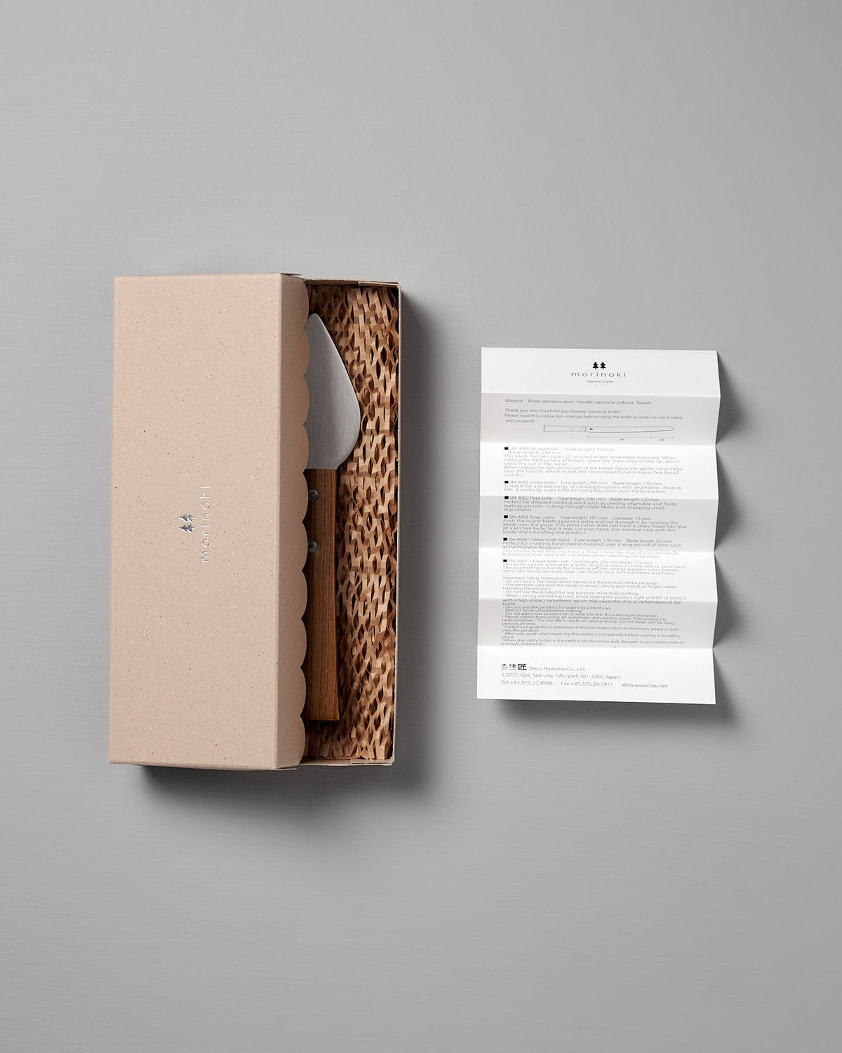 A beige box with scalloped edges holds the Morinoki Hard Cheese Knife by Shizu Hamono Co., featuring a stainless steel blade on textured padding, beside a folded instruction sheet on a light gray surface.