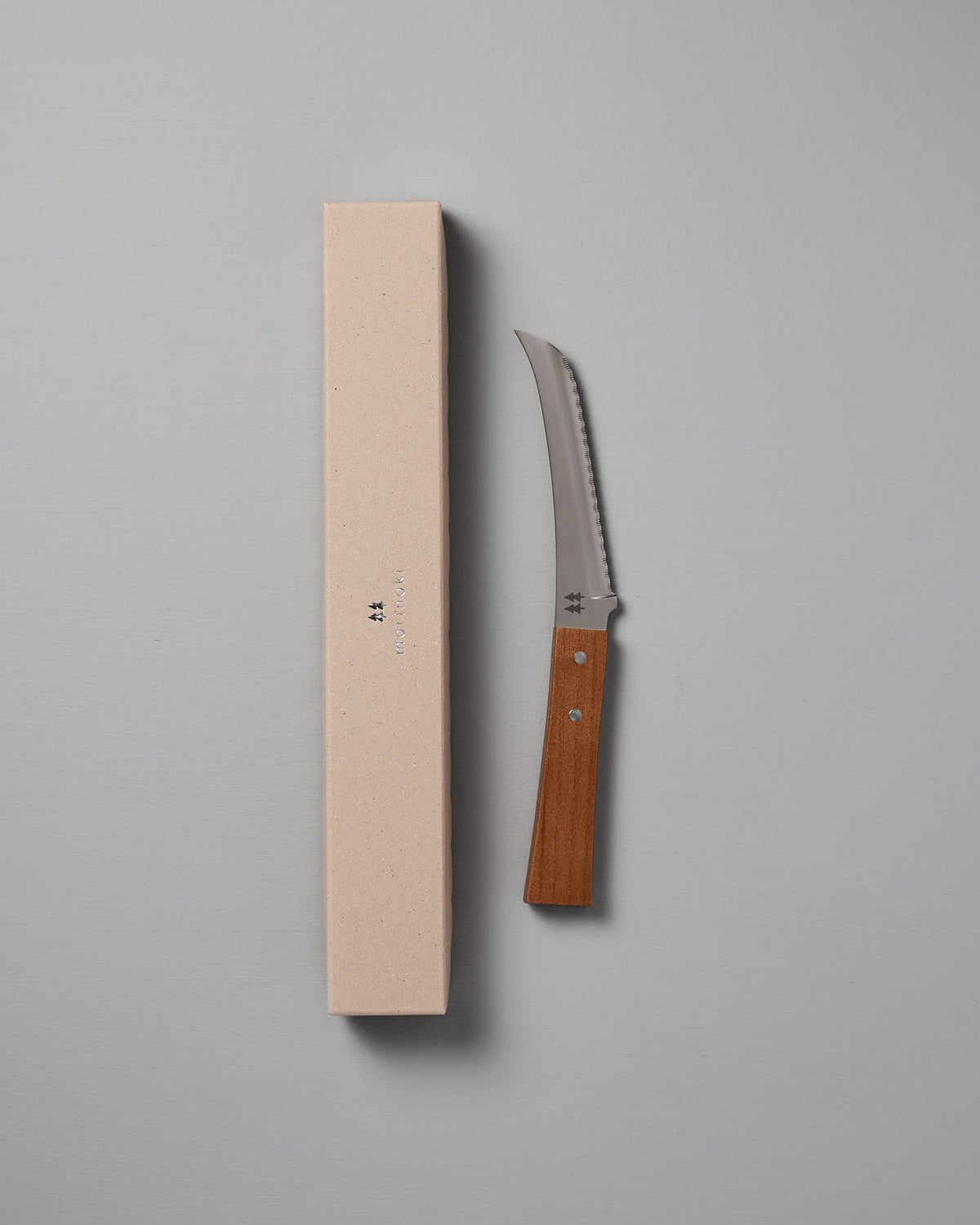 The Morinoki Soft Cheese Knife by Shizu Hamono Co., featuring a Japanese Zelkova handle, rests beside a beige rectangular box on a light gray surface.