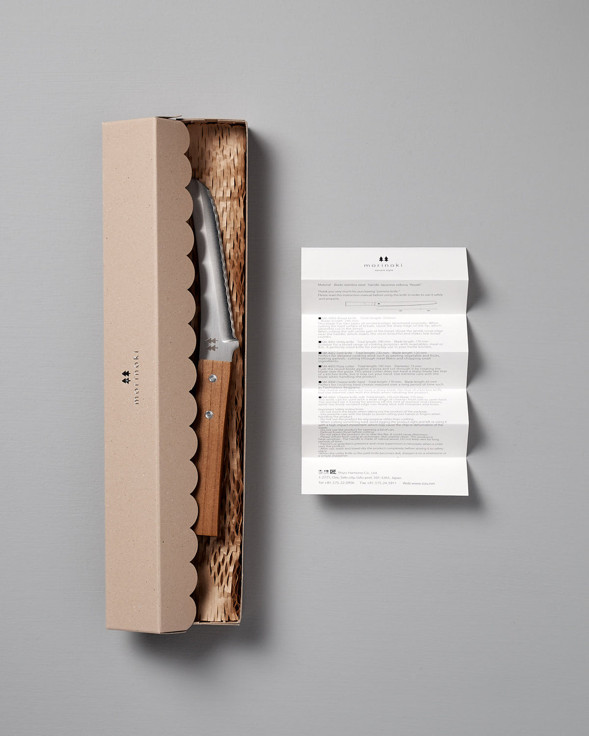 The Morinoki Soft Cheese Knife by Shizu Hamono Co. features a Japanese Zelkova wood handle and is presented in a cardboard box with textured packing and an instruction leaflet, highlighting exquisite Japanese craftsmanship.
