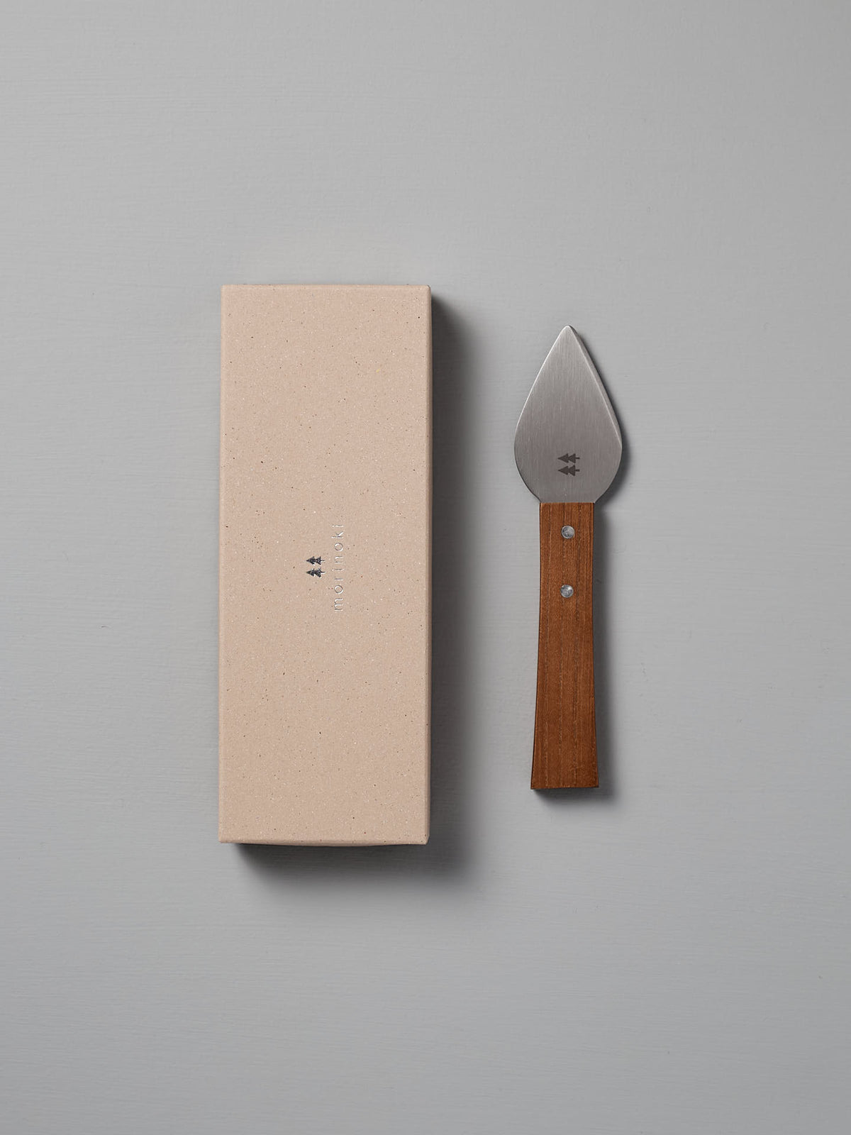 The Morinoki Hard Cheese Knife by Shizu Hamono Co., featuring a wide stainless steel blade and wooden handle, is placed next to a closed beige box on a light gray surface.