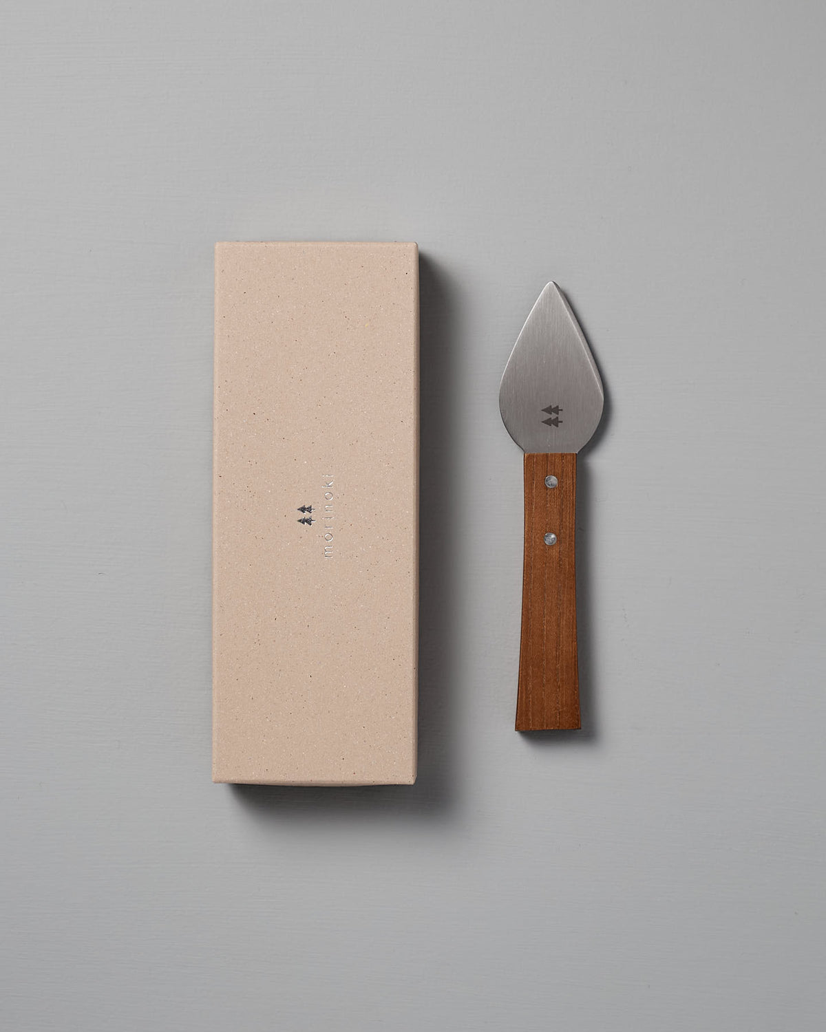 The Morinoki Hard Cheese Knife by Shizu Hamono Co., featuring a wide stainless steel blade and wooden handle, is placed next to a closed beige box on a light gray surface.