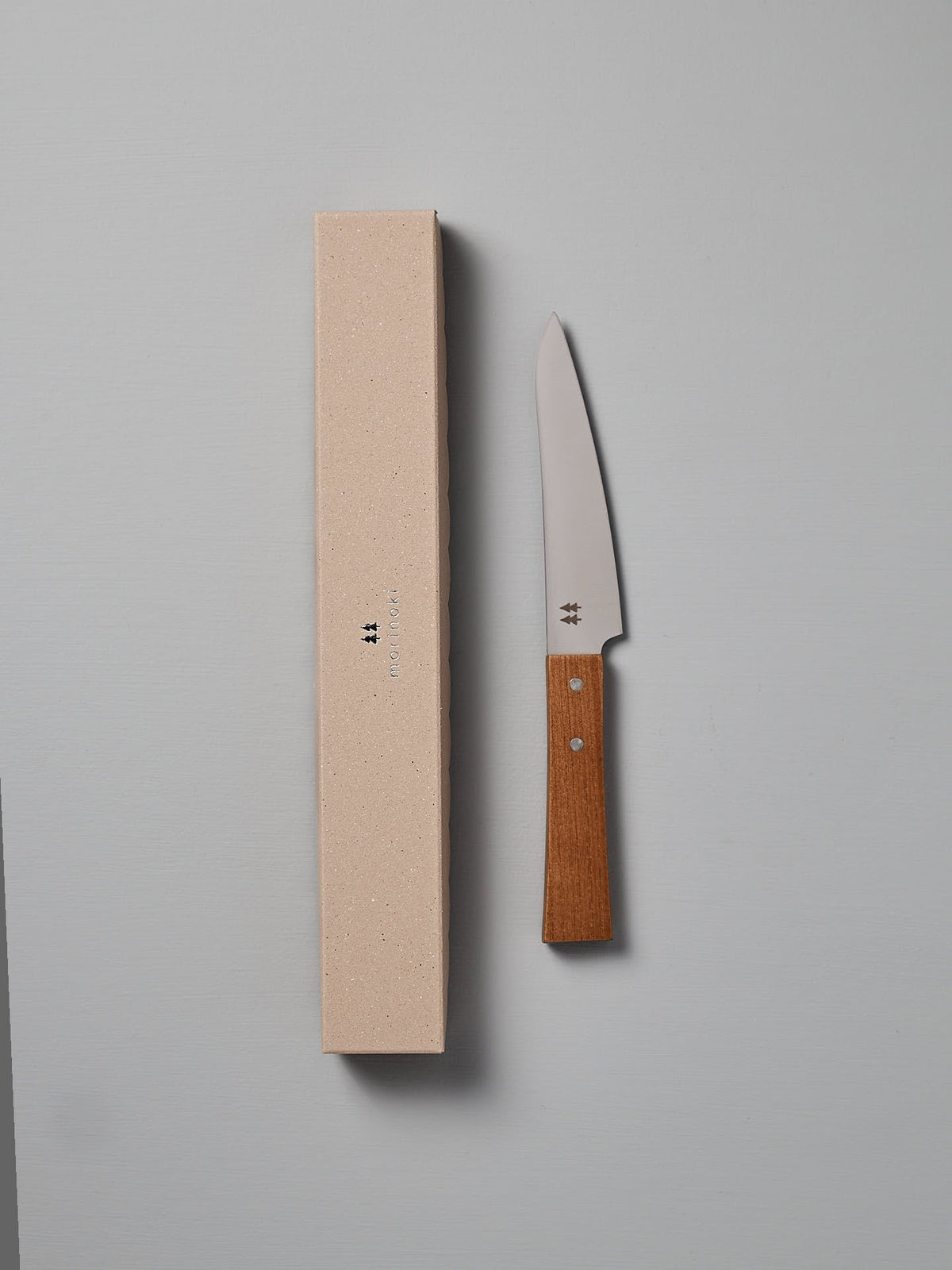 A Morinoki Petite Knife by Shizu Hamono Co., featuring a Japanese Zelkova wood handle, is placed alongside its beige, rectangular box on a plain white surface.