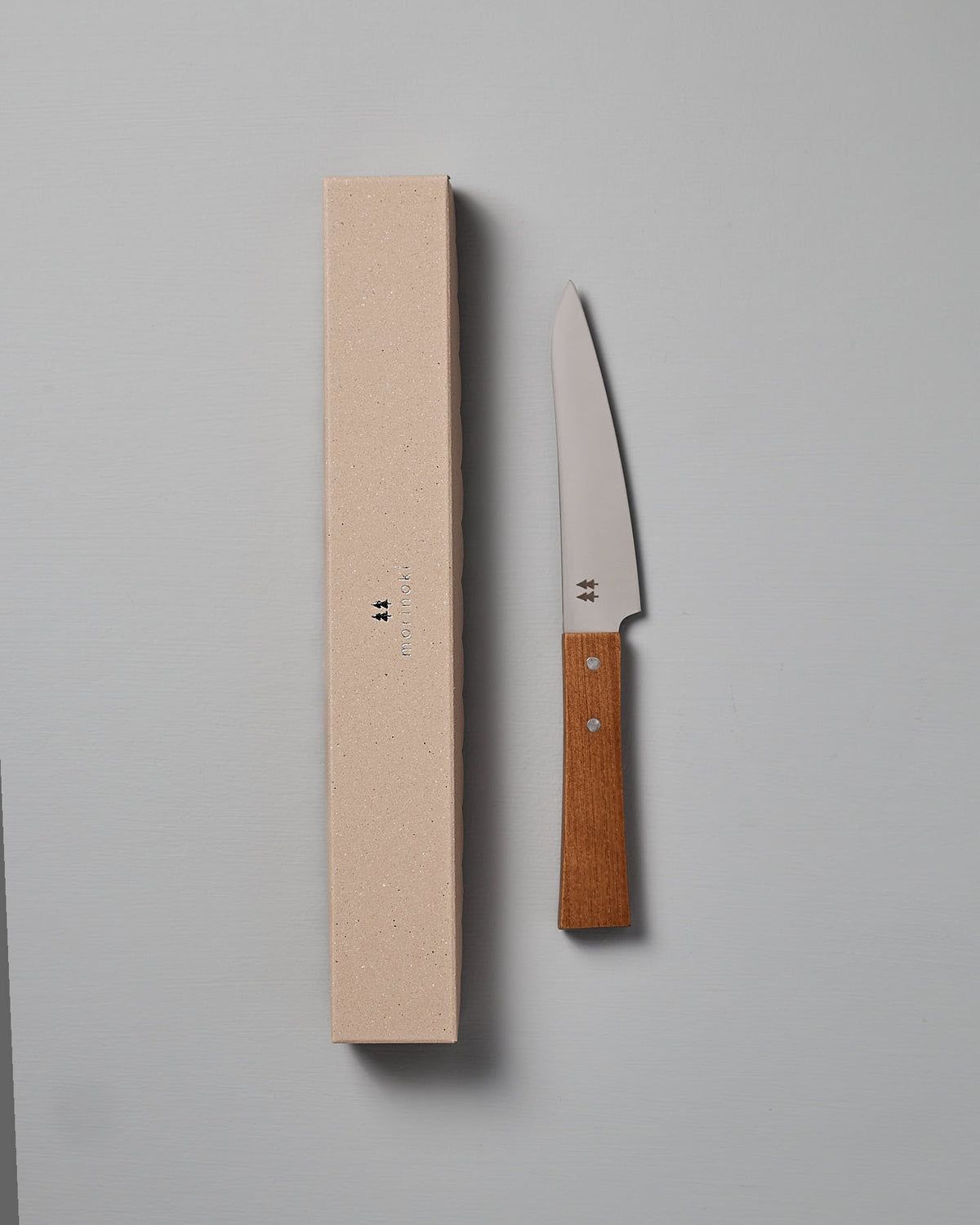 The Morinoki Petite Knife by Shizu Hamono Co., featuring a stainless steel blade and wooden handle, is elegantly displayed beside its beige box on a gray surface.