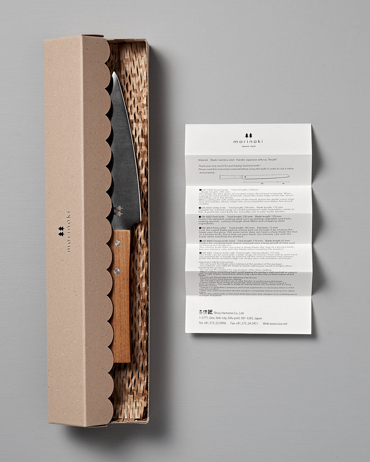 The Morinoki Petite Knife by Shizu Hamono Co. features an ergonomic wooden handle and a sharp stainless steel blade for precise kitchen tasks. It rests in a textured cardboard box with an instruction leaflet on a gray background.