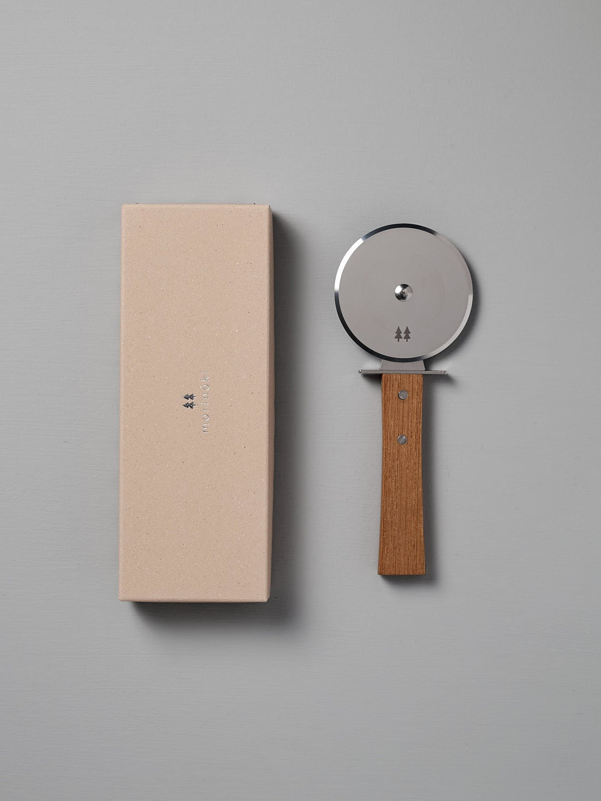 The Morinoki Pizza Wheel by Shizu Hamono Co., featuring a stainless steel blade and a Japanese Zelkova handle, is placed next to a beige rectangular box on a gray surface.