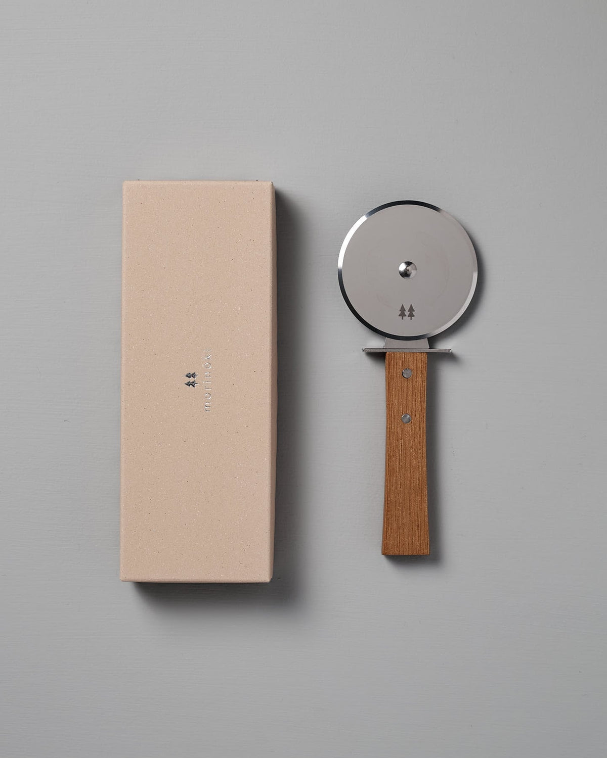 The Morinoki Pizza Wheel by Shizu Hamono Co., featuring a stainless steel blade and a Japanese Zelkova handle, is placed next to a beige rectangular box on a gray surface.