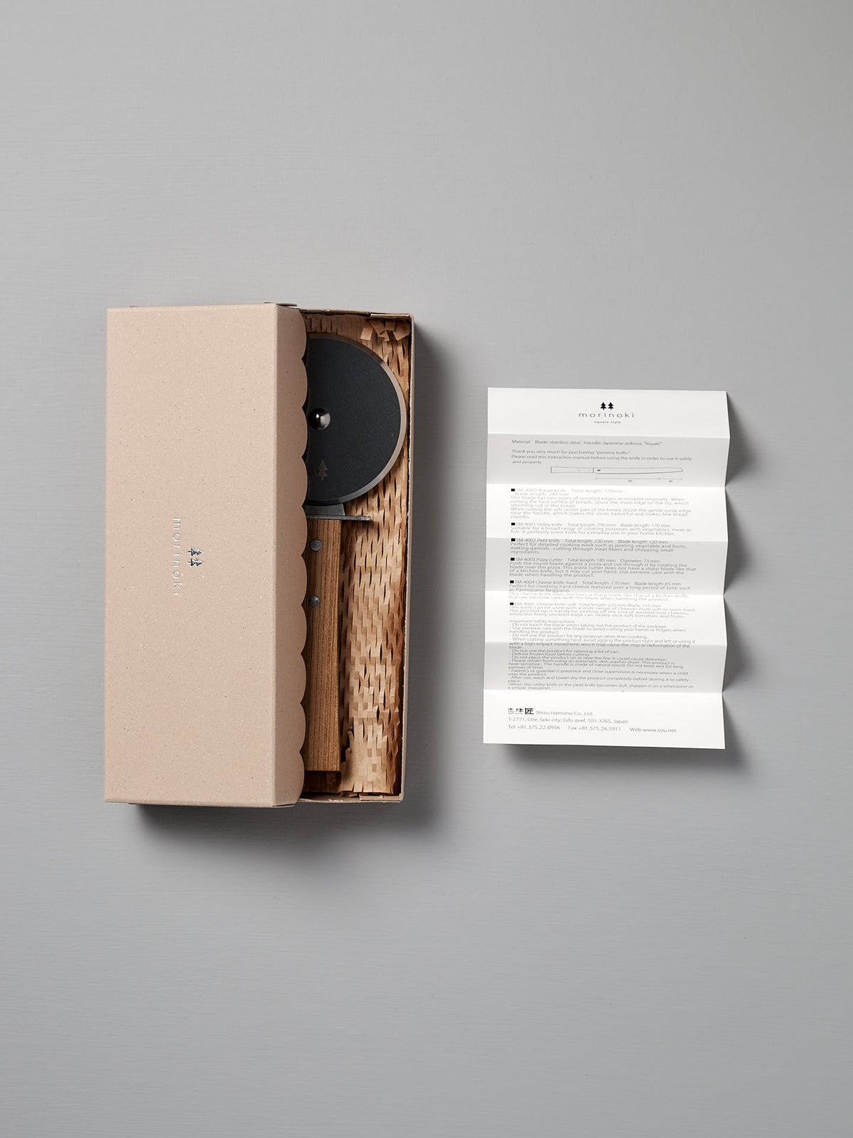 A scalloped-edge cardboard box holds the Shizu Hamono Co. Morinoki Pizza Wheel with a Japanese Zelkova handle; an unfolded instruction sheet is positioned to the right on a gray background.