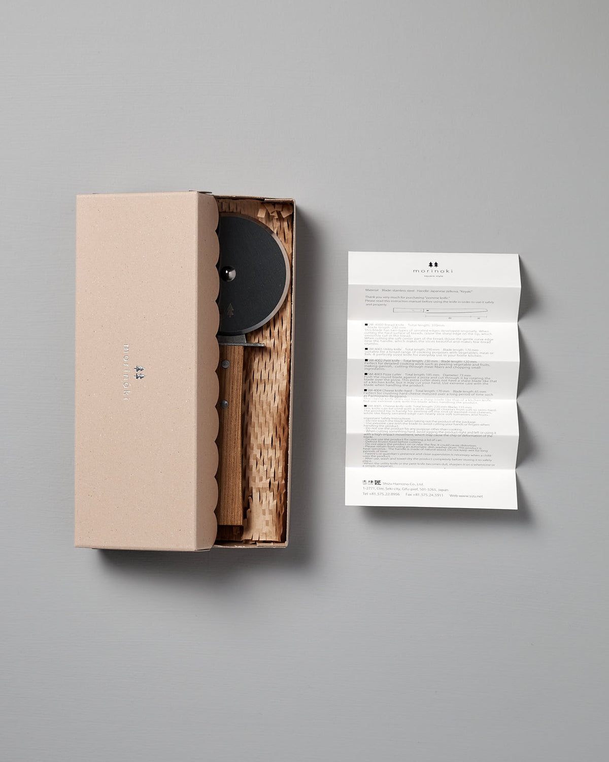 A scalloped-edge cardboard box holds the Shizu Hamono Co. Morinoki Pizza Wheel with a Japanese Zelkova handle; an unfolded instruction sheet is positioned to the right on a gray background.