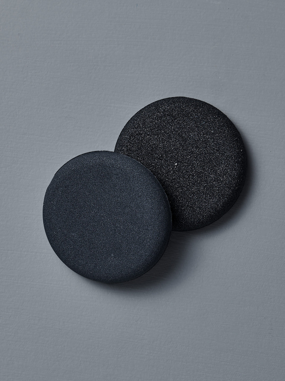 Two round, black Dual-Sided Water Stones from The Knife Sharpener sit side by side on a sleek gray surface, ideal as compact portable sharpeners.