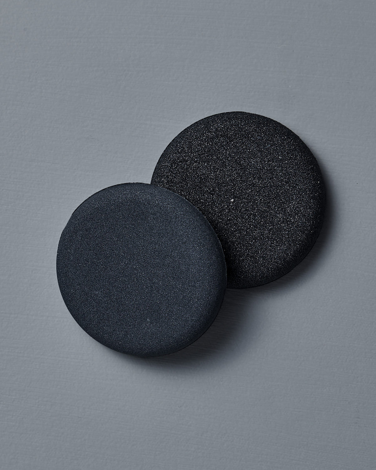 Two round, black Dual-Sided Water Stones from The Knife Sharpener sit side by side on a sleek gray surface, ideal as compact portable sharpeners.