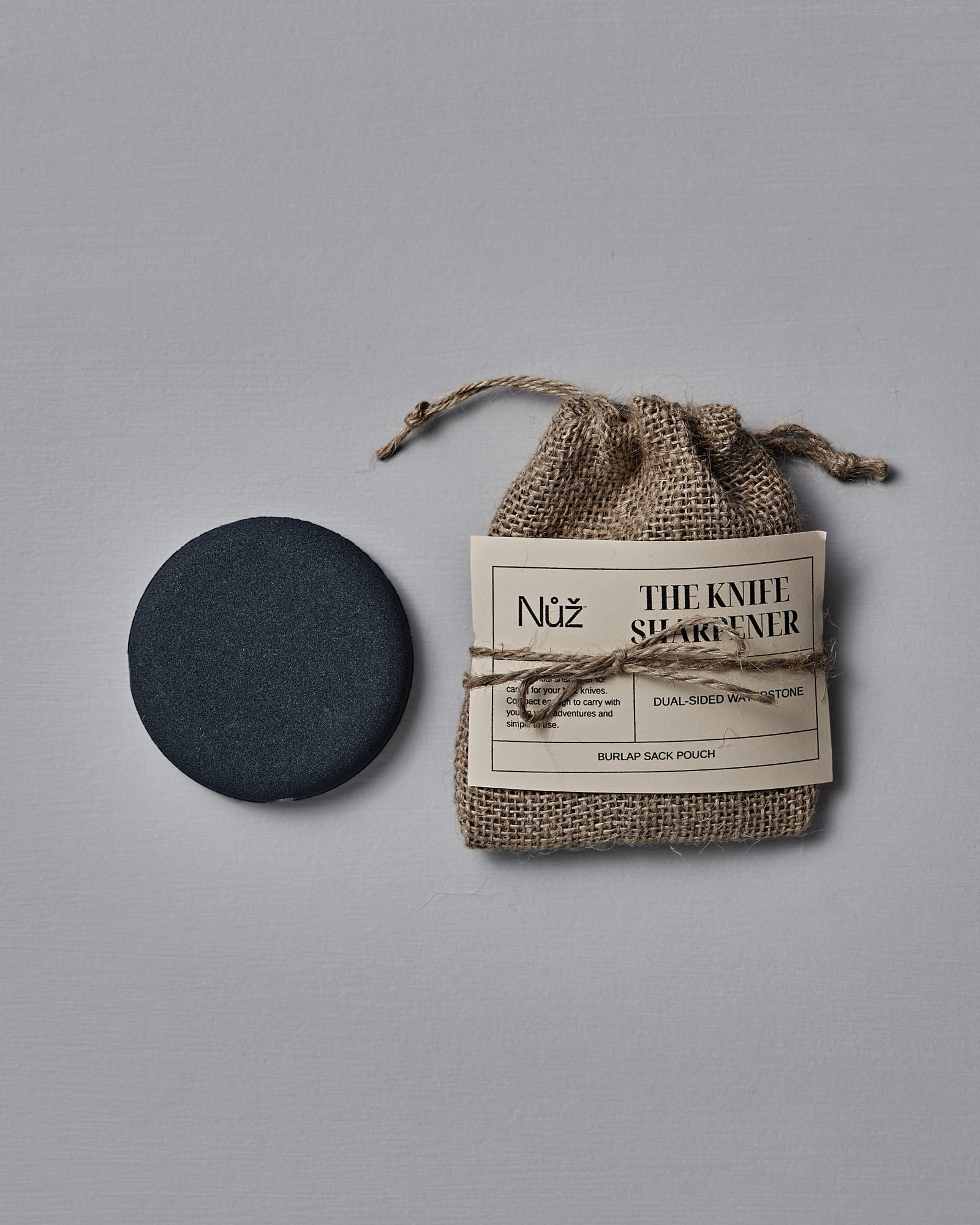 The Knife Sharpener – Dual-Sided Water Stone rests next to a burlap pouch labeled "The Knife Sharpener" on a gray surface.