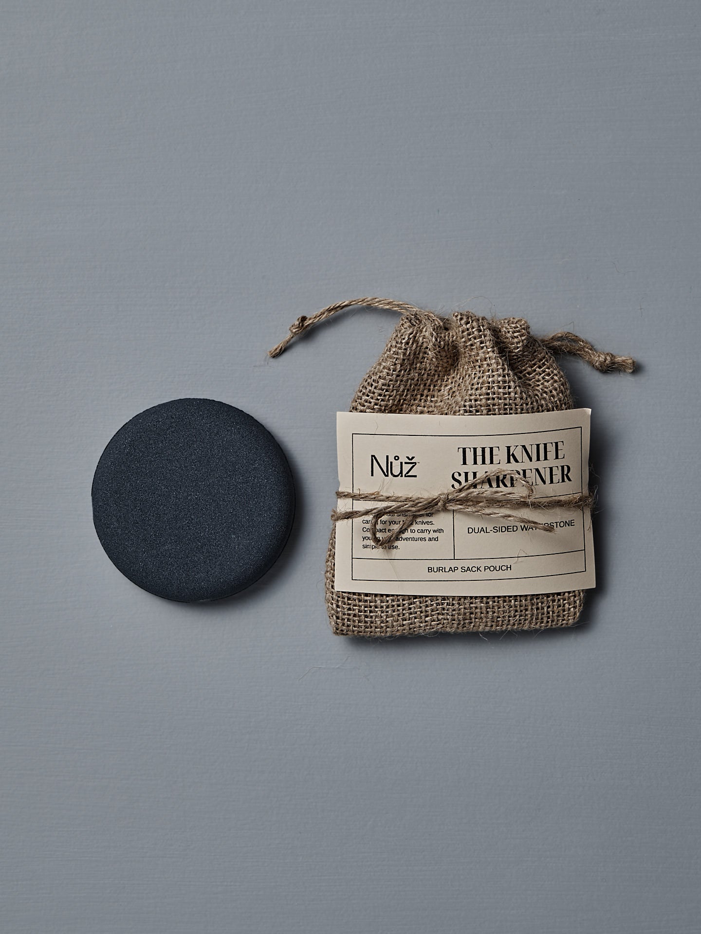 The Knife Sharpener – Dual-Sided Water Stone rests next to a burlap pouch labeled "The Knife Sharpener" on a gray surface.