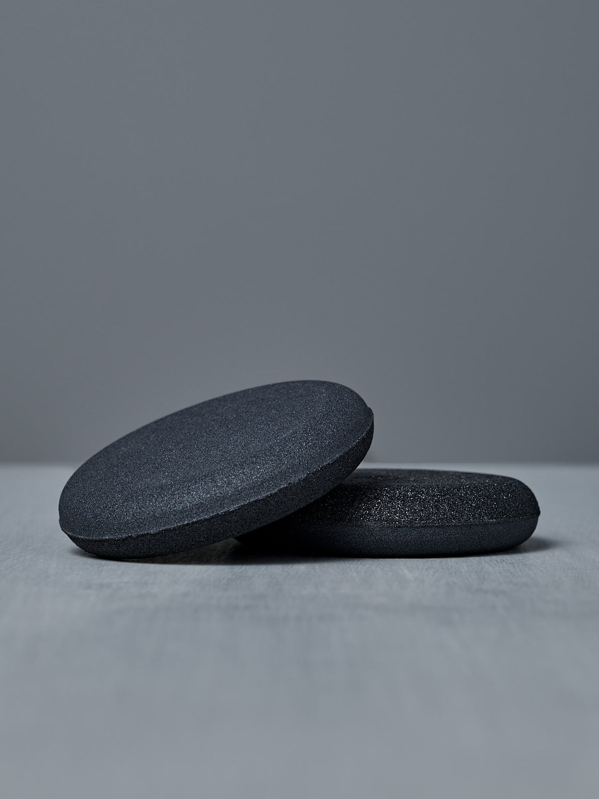 Two smooth, round, dark gray “The Knife Sharpener – Dual-Sided Water Stone” stones rest on a light gray surface against a plain gray background.