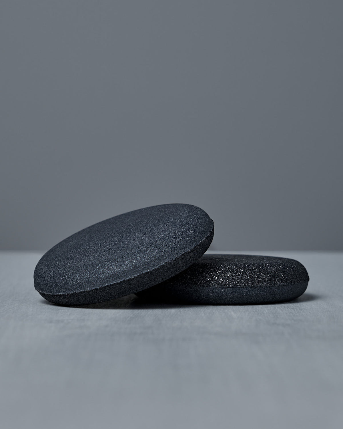 Two smooth, round, dark gray “The Knife Sharpener – Dual-Sided Water Stone” stones rest on a light gray surface against a plain gray background.