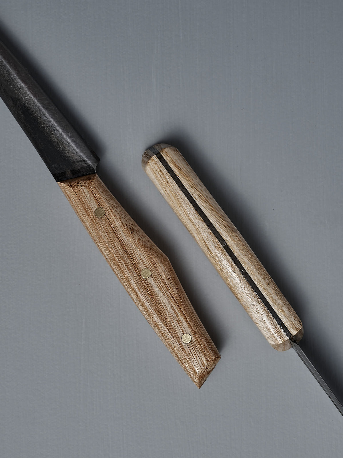 Two №1 – Spread Knives with handcrafted wooden handles rest on a gray surface—one displays the blade and handle, while the other shows the handle and spine of its Swedish carbon steel blade.