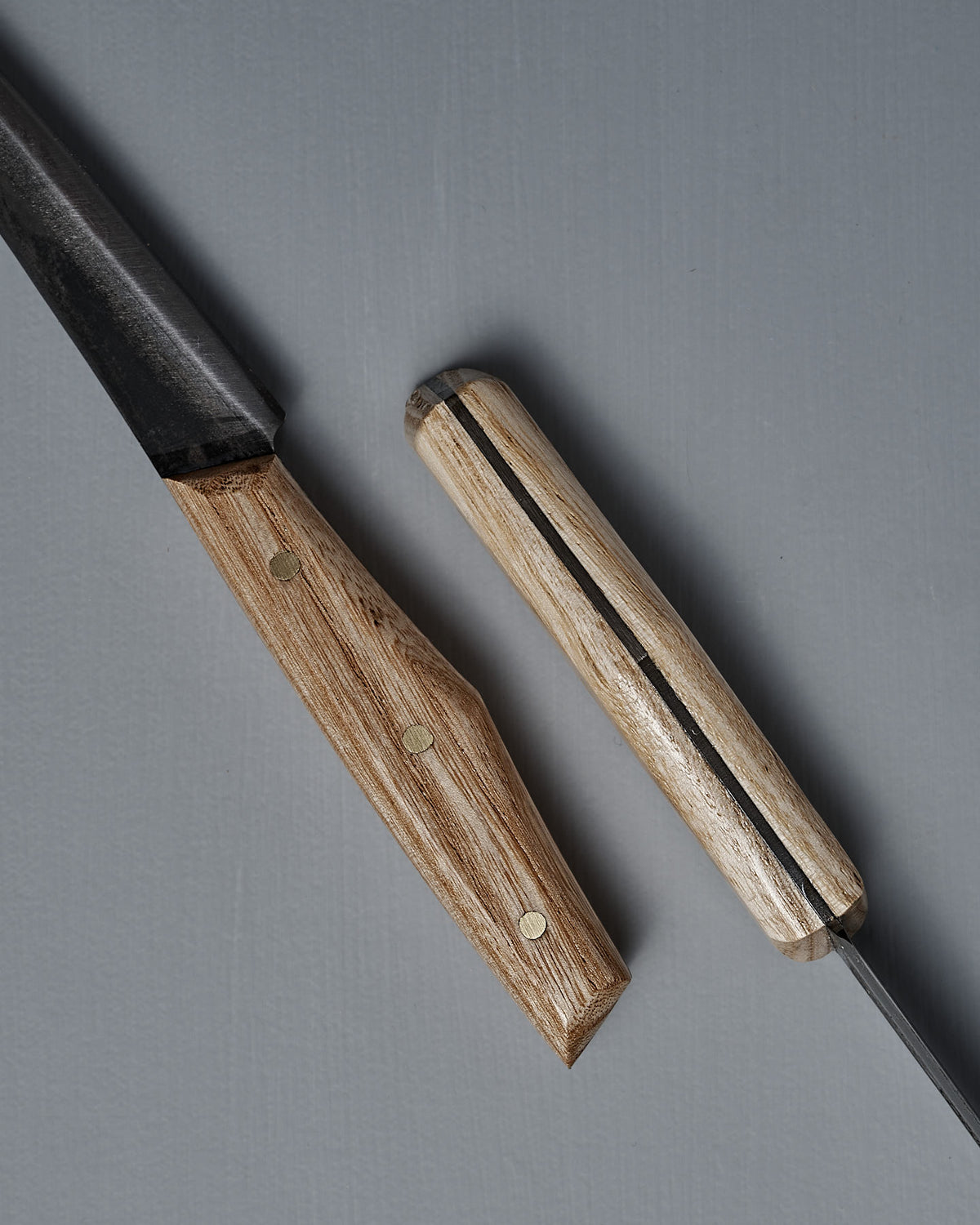 Two №1 – Spread Knives with handcrafted wooden handles rest on a gray surface—one displays the blade and handle, while the other shows the handle and spine of its Swedish carbon steel blade.