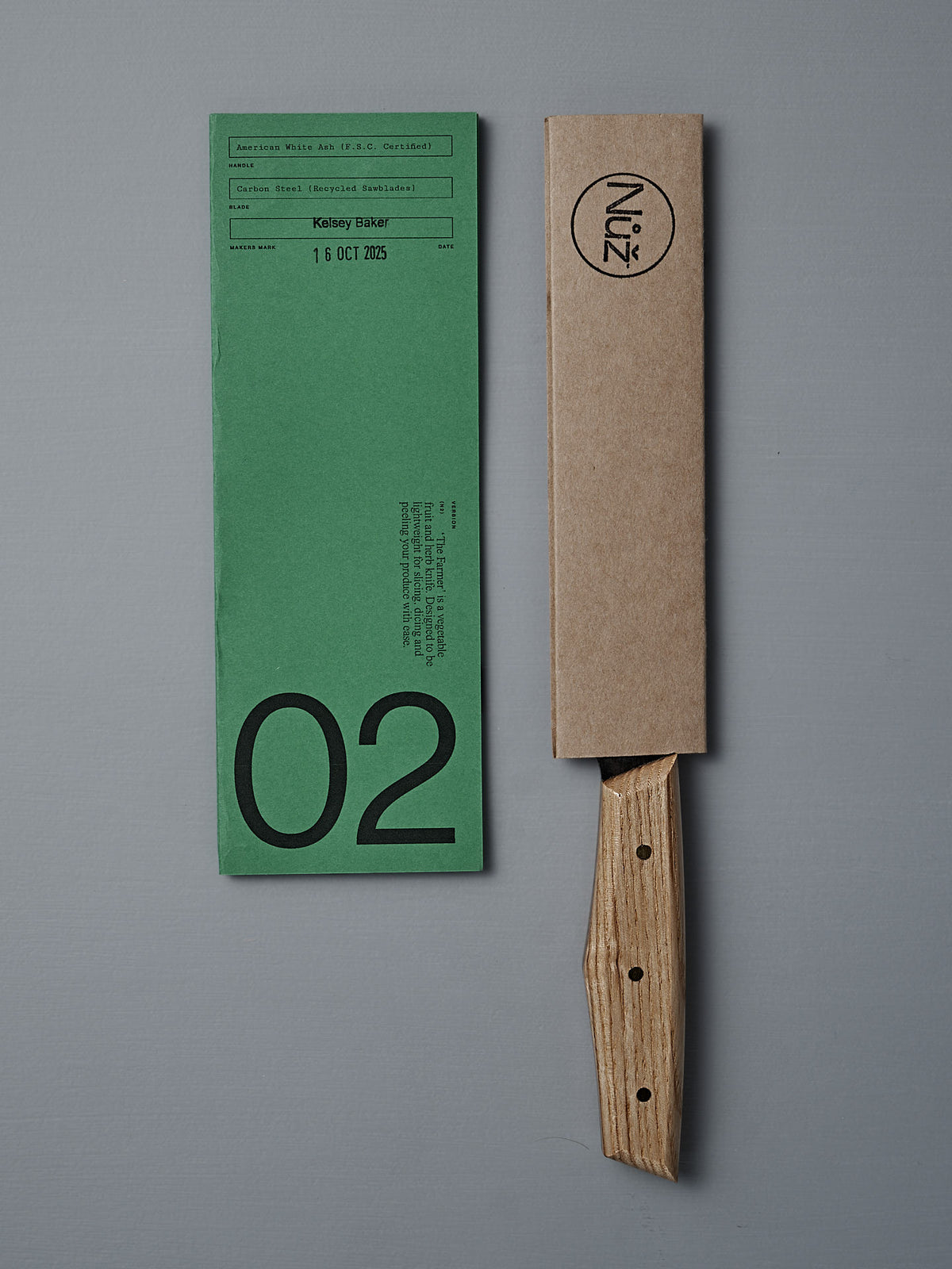A green paper form labeled "02" sits beside the №2 – Farmer Knife with an American Ash handle, partly sheathed in brown cardboard bearing "Núz" branding, all arranged on a gray surface.