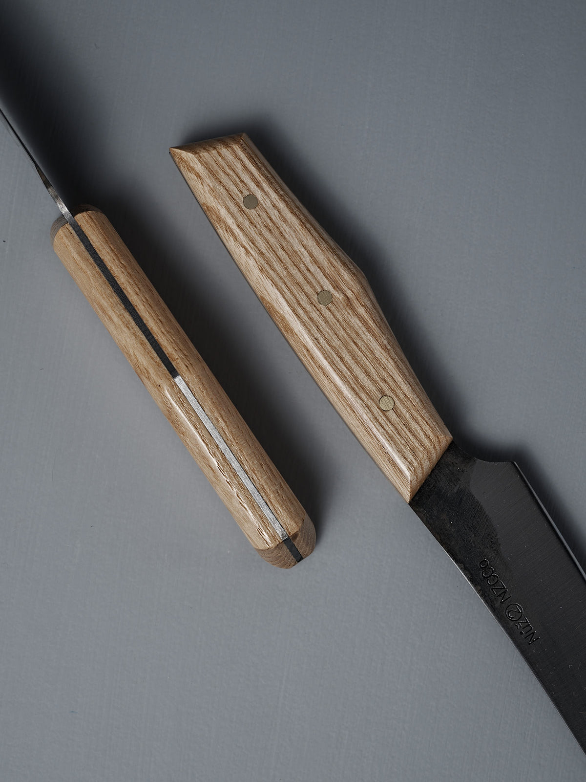 Two №2 – Farmer Knives with American Ash handles rest on a flat, gray surface. Their handcrafted handles are shown up close, highlighting the unique shapes and natural wood grain patterns.