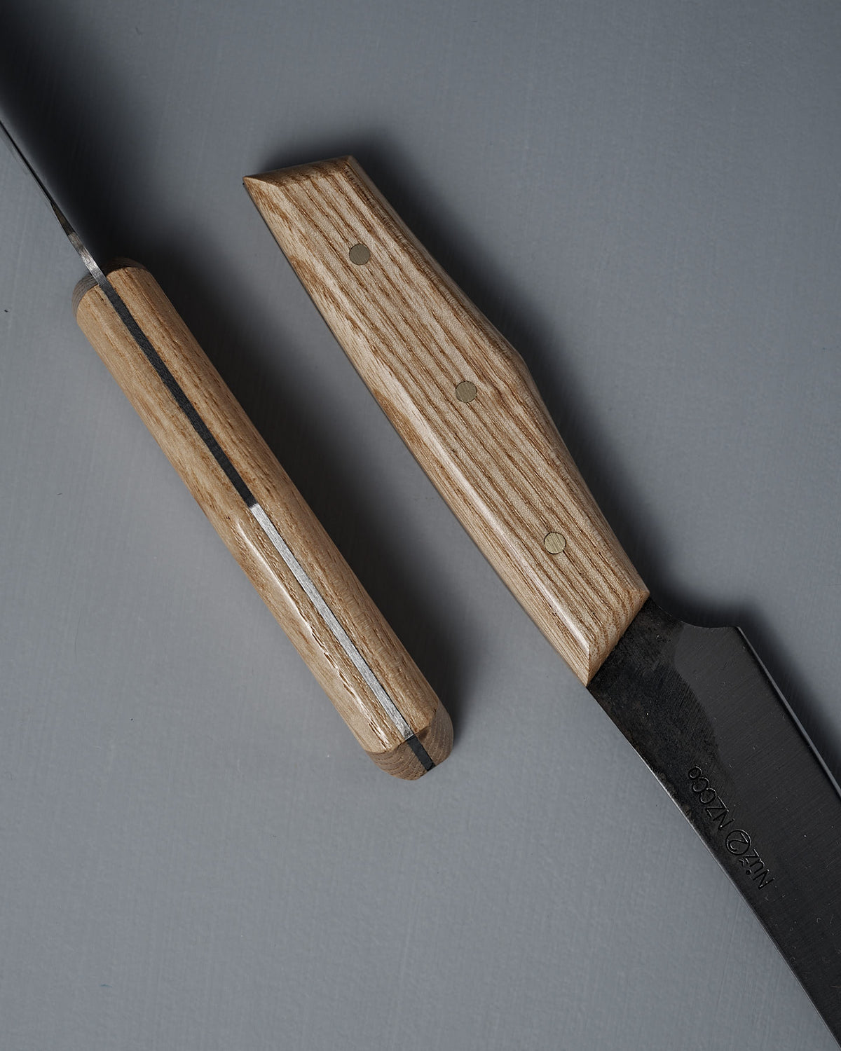 Two №2 – Farmer Knives with American Ash handles rest on a flat, gray surface. Their handcrafted handles are shown up close, highlighting the unique shapes and natural wood grain patterns.