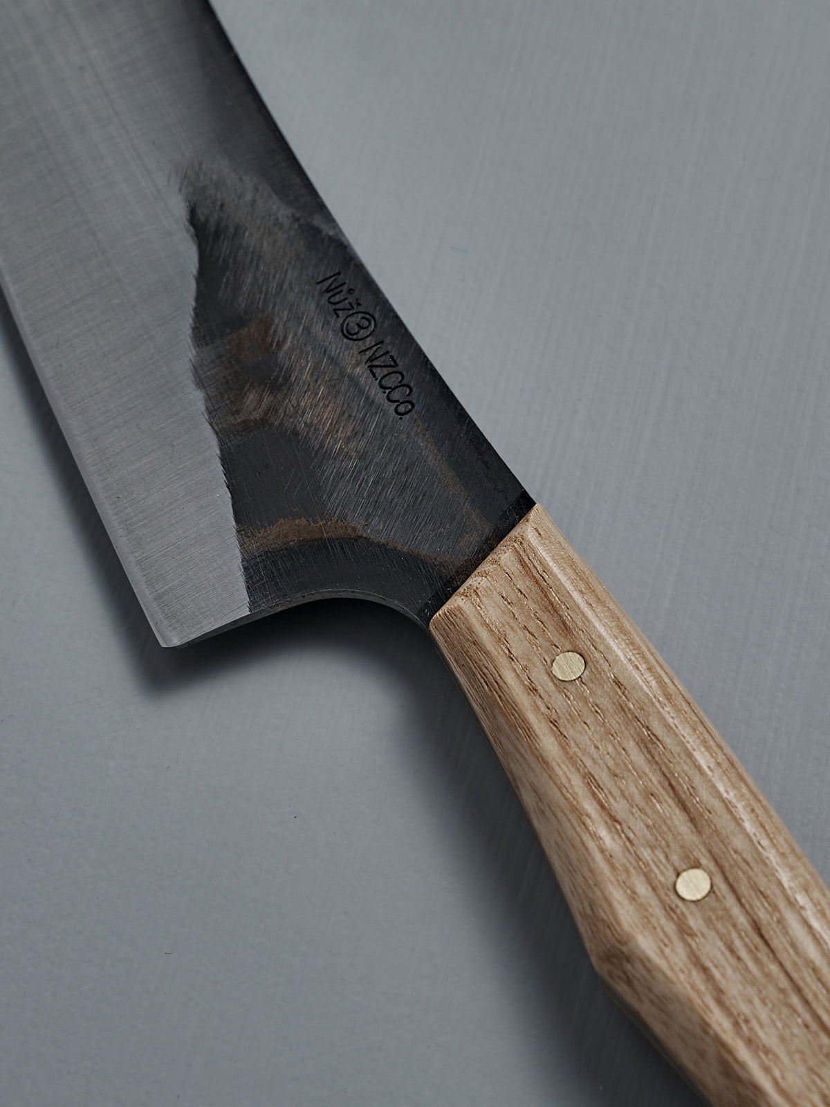 A close-up of the №3 – Daily Knife, showcasing its American Ash handle and partially polished carbon steel blade, resting on a gray surface.