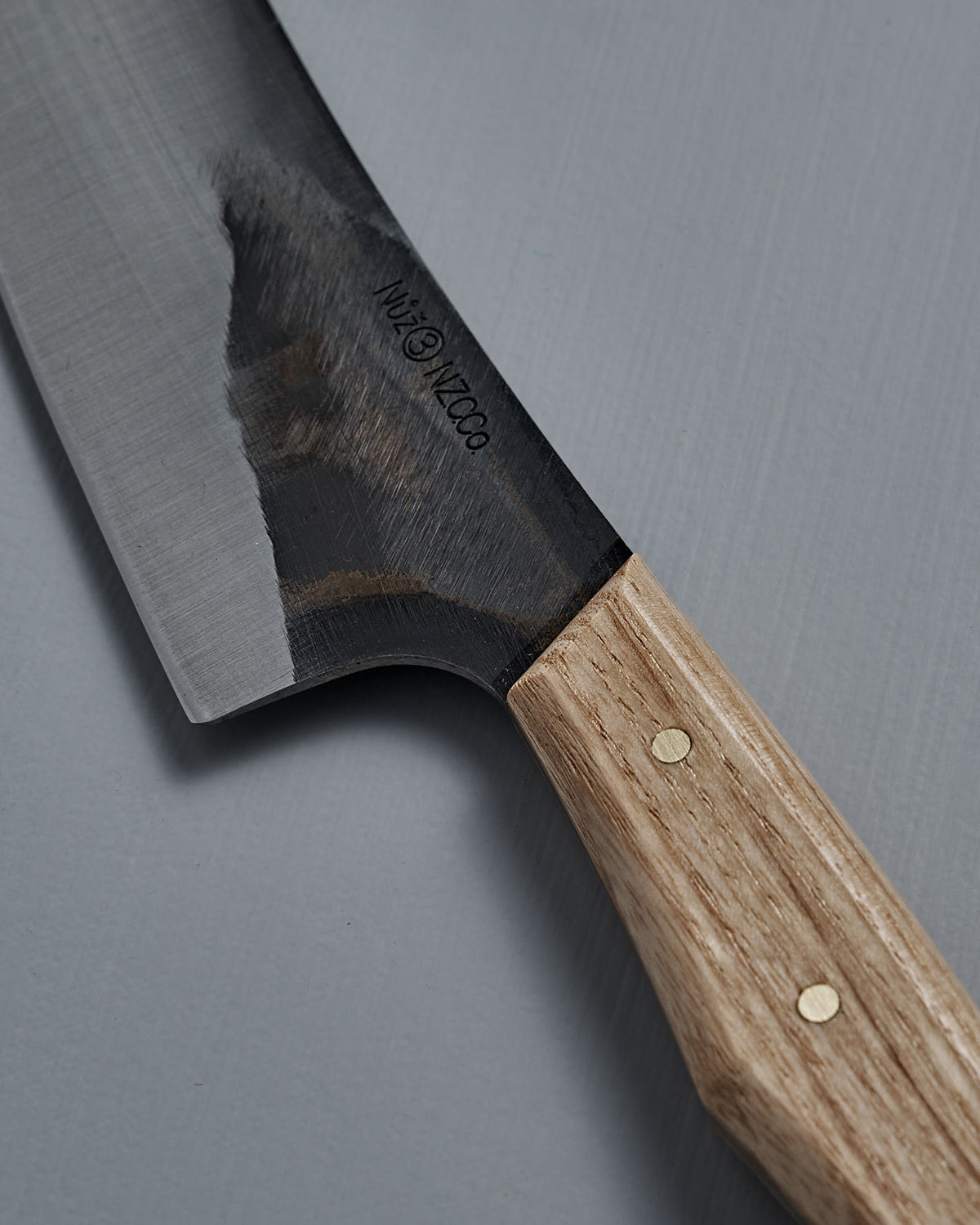 A close-up of the №3 – Daily Knife, showcasing its American Ash handle and partially polished carbon steel blade, resting on a gray surface.