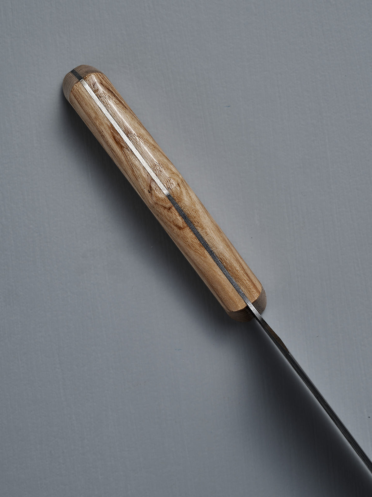 A top-down close-up of the №3 – Daily Knife, featuring a handcrafted American Ash handle and carbon steel blade, displayed on a gray surface.