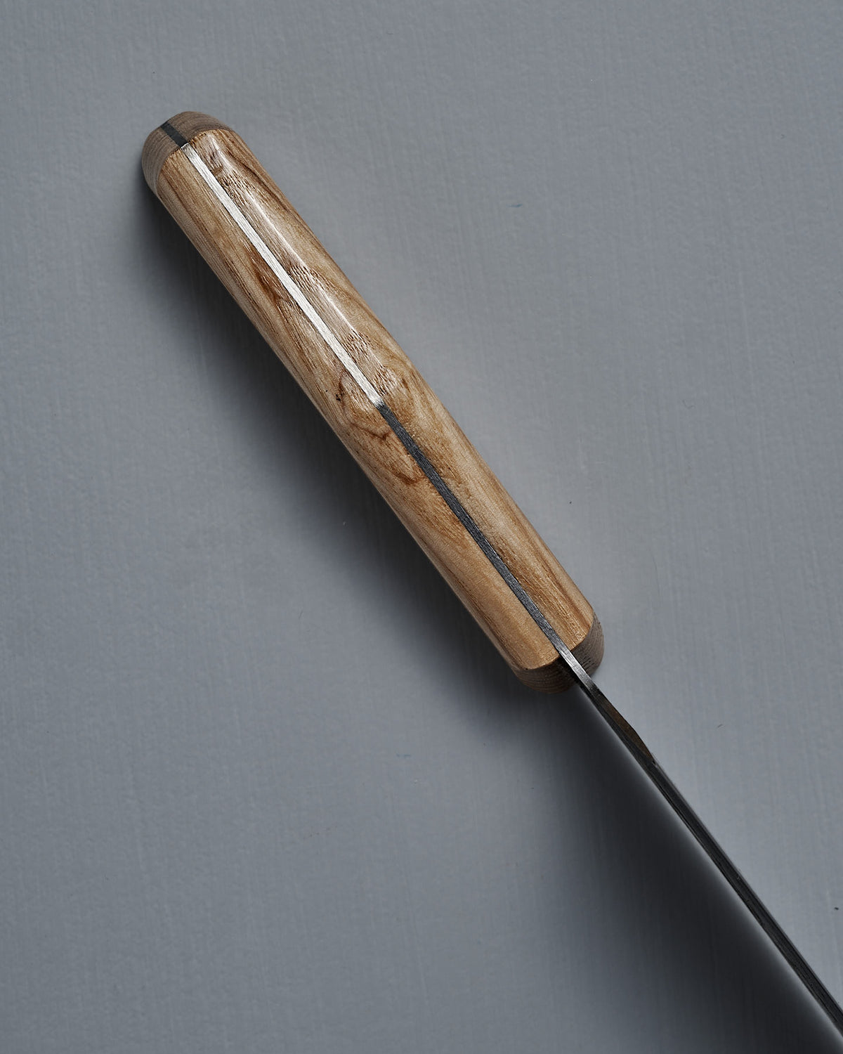 A top-down close-up of the №3 – Daily Knife, featuring a handcrafted American Ash handle and carbon steel blade, displayed on a gray surface.