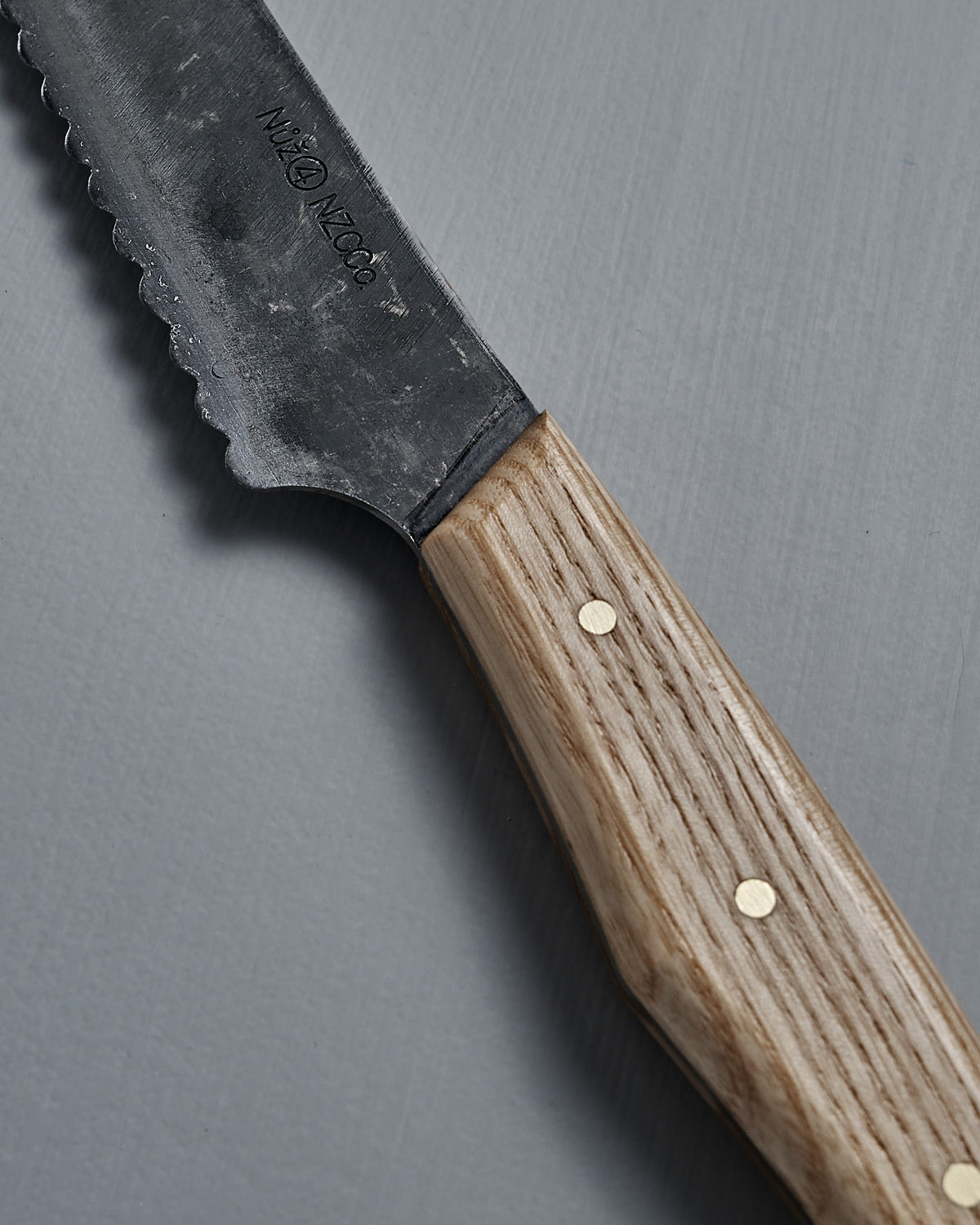 The №4 – Baker Knife, a handcrafted New Zealand bread knife with a carbon steel blade and wooden handle, rests on a gray surface.