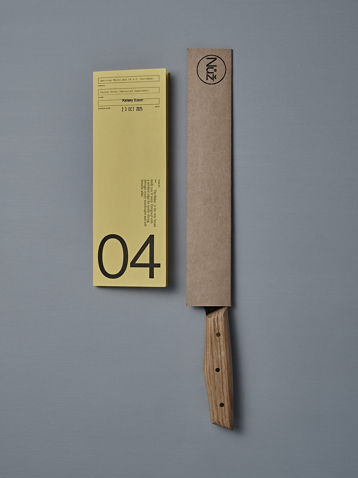 The №4 – Baker Knife, a handcrafted New Zealand bread knife with a wooden handle and carbon steel blade, partially sheathed in cardboard, rests beside a yellow “04” label on a gray surface.
