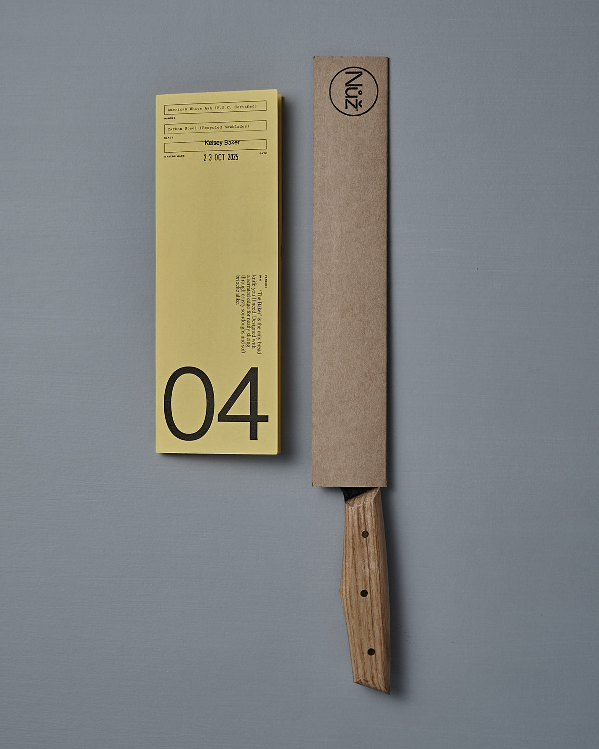 The №4 – Baker Knife, a handcrafted New Zealand bread knife with a wooden handle and carbon steel blade, partially sheathed in cardboard, rests beside a yellow “04” label on a gray surface.