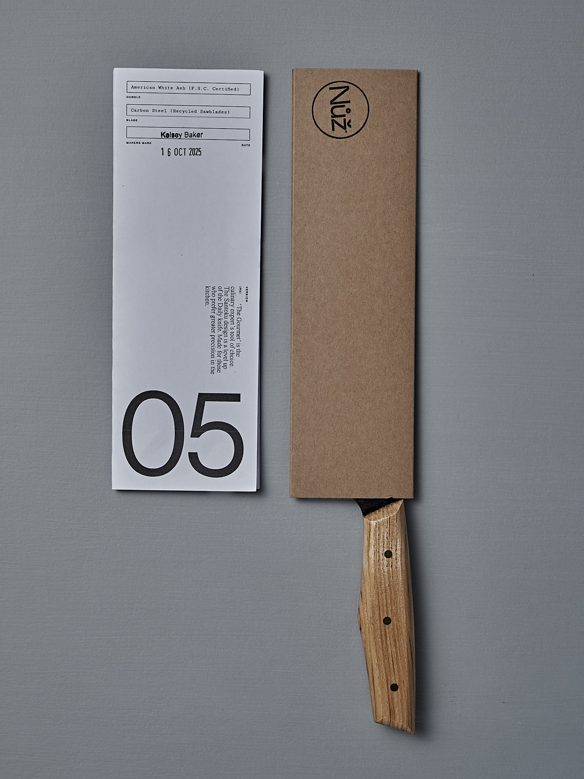 The №5 – Gourmet Knife, featuring a handcrafted wooden handle and a brown cardboard sheath, rests beside a white paper slip marked with a bold "05".