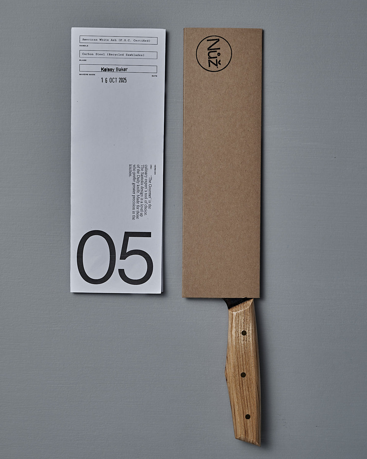 The №5 – Gourmet Knife, featuring a handcrafted wooden handle and a brown cardboard sheath, rests beside a white paper slip marked with a bold "05".