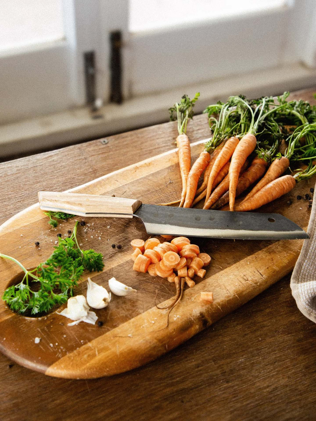 The №5 – Gourmet Knife rests on a wooden cutting board alongside sliced carrots, whole carrots with greens, garlic cloves, parsley, and peppercorns on a wooden table.