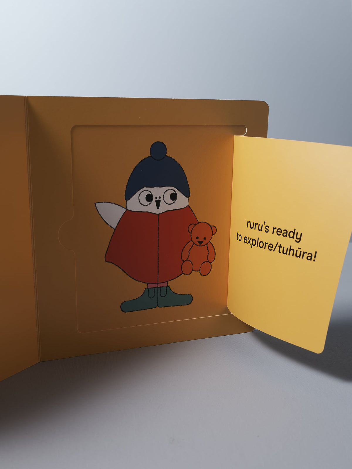 A Let&#39;s Go Ruru - by Kate Muir, an owl with a beanie and glasses, holding a teddy bear, depicted on a foldable card with the phrase &quot;Ruru&#39;s ready for an