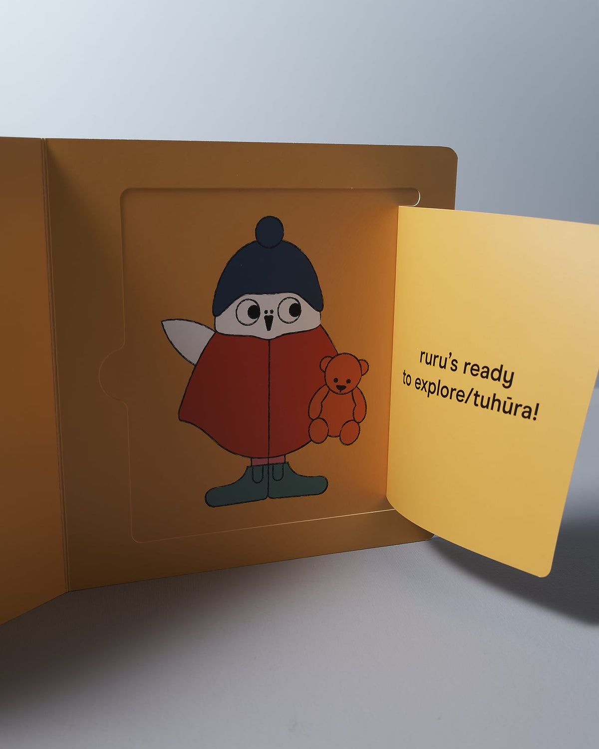 A Let's Go Ruru - by Kate Muir, an owl with a beanie and glasses, holding a teddy bear, depicted on a foldable card with the phrase "Ruru's ready for an