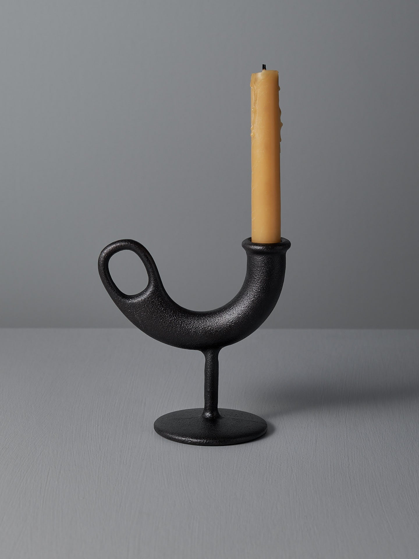 The Ildhane Candle-stick Holder by Nedre Foss is a modernist black, curved metal piece featuring a circular base, elegantly displaying a tall, unlit yellow candle against a plain gray background.