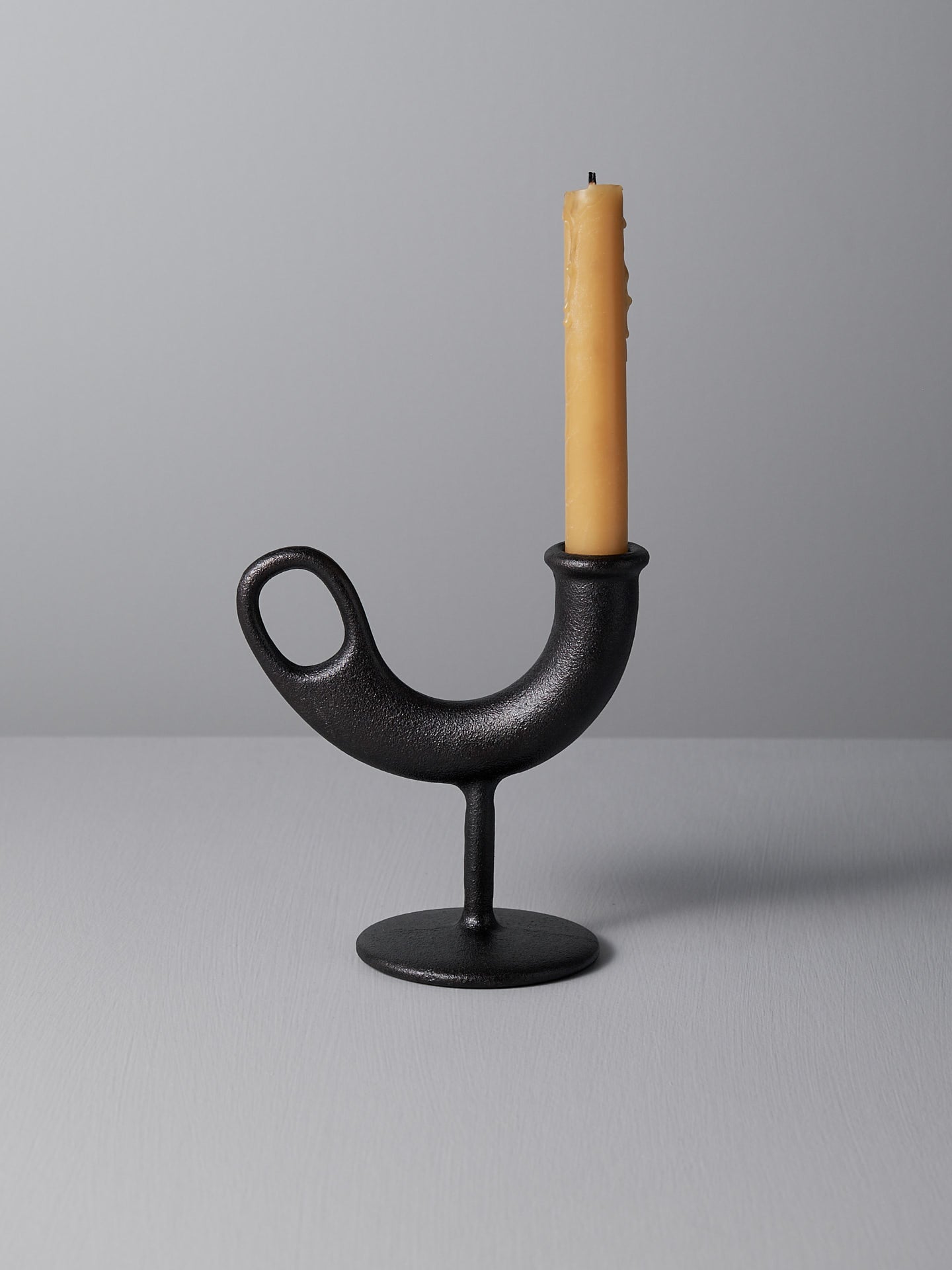 A uniquely shaped black Ildhane Candlestick Holder by Nedre Foss, crafted by designers Anderssen & Voll, supports a partially burned yellow candle as it sits on a light gray surface against a matching gray background, evoking the essence of a modernist landscape.