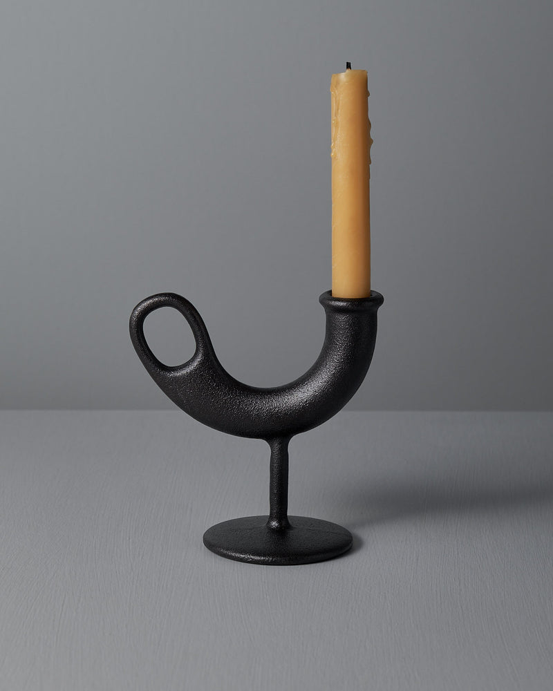 The Ildhane Candle-stick Holder by Nedre Foss is a modernist black, curved metal piece featuring a circular base, elegantly displaying a tall, unlit yellow candle against a plain gray background.