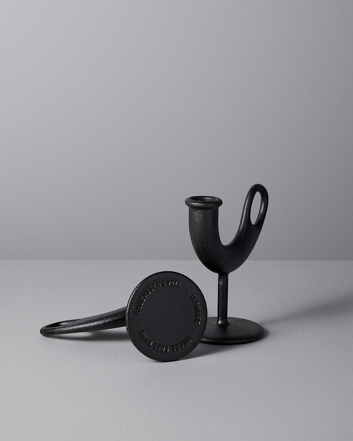 Two black Ildhane candle-stick holders from Nedre Foss, designed by Anderssen & Voll, enhance the scene with one standing upright and the other gracefully lying on a gray surface against a matching backdrop, exuding a distinct modernist charm.