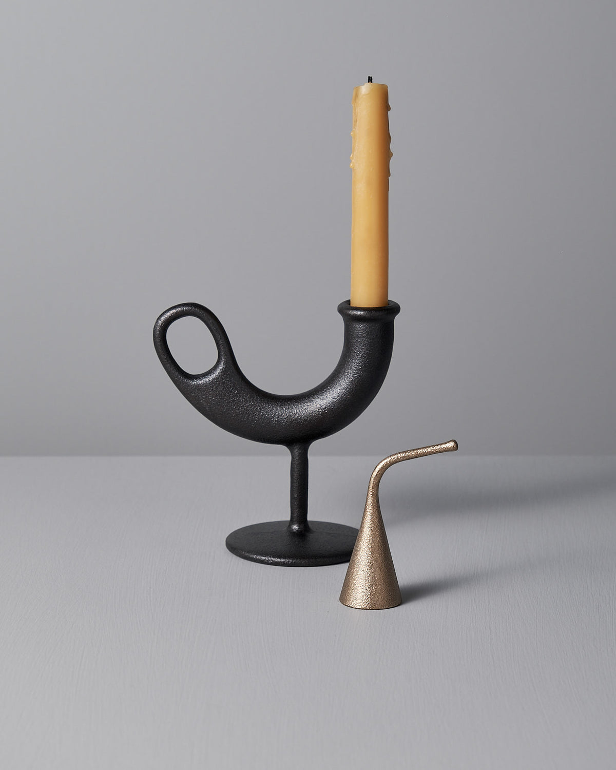 A black metal candle holder with a yellow candle and the small Sammu Candle Snuffer by Nedre Foss on a gray surface against a gray background.