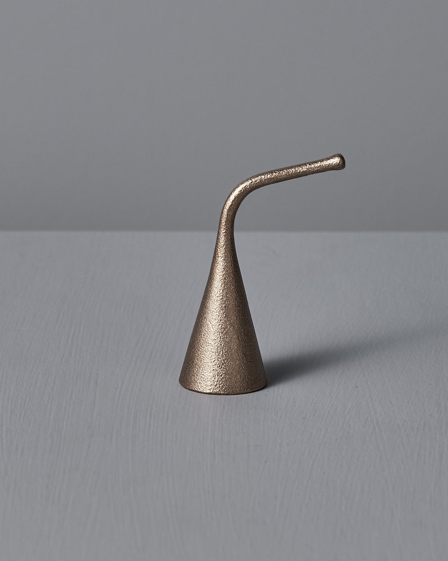 A Sammu Candle Snuffer by Nedre Foss, featuring a brass-colored, conical design with a curved handle, is placed on a gray surface against a light gray background, evoking traditional Finnish design.